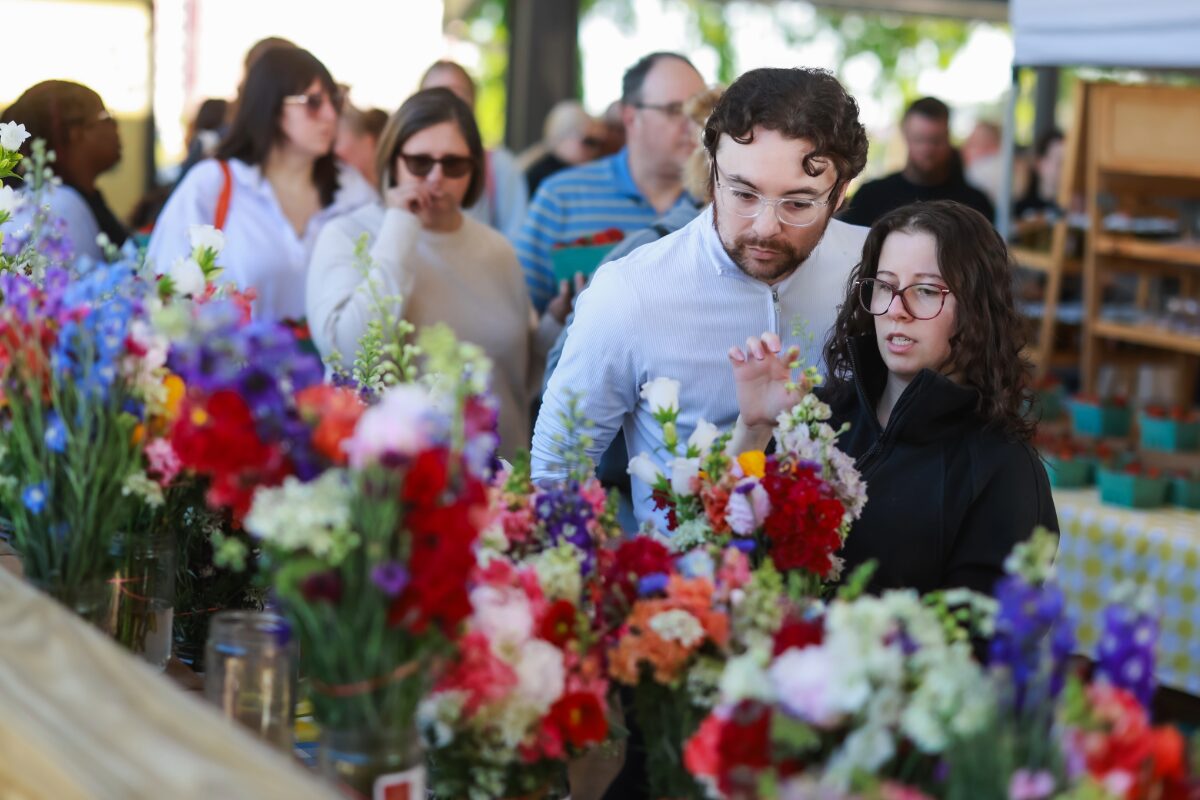Couple stands at a flower stall, examining bright bouquets of red, pink, and purple flowers while other shoppers browse in the background.