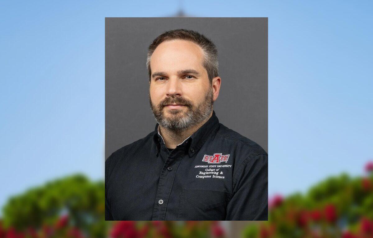 Portrait of a man with short hair and a gray beard, wearing a black Arkansas State University Engineering & Computer Science shirt against a gray backdrop.