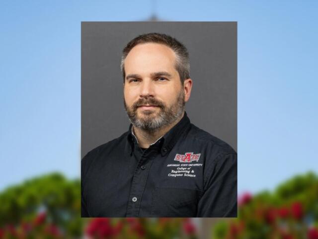 Portrait of a man with short hair and a gray beard, wearing a black Arkansas State University Engineering & Computer Science shirt against a gray backdrop.