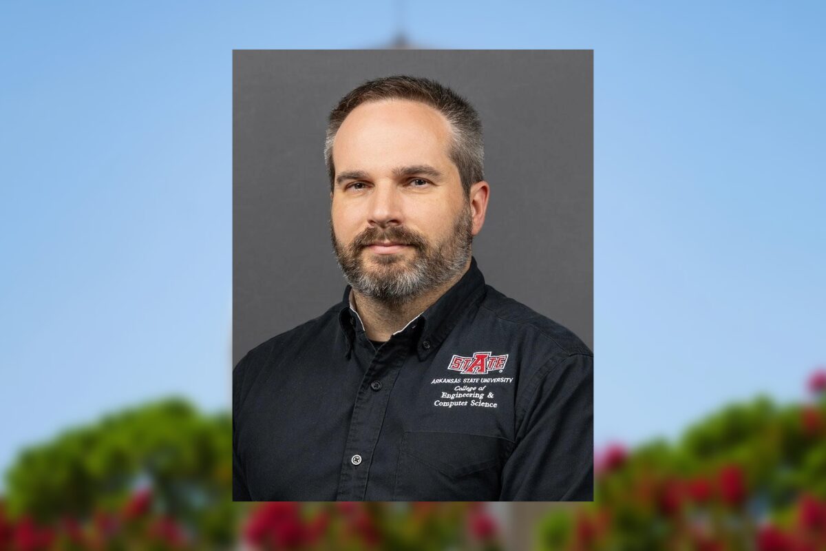 Portrait of a man with short hair and a gray beard, wearing a black Arkansas State University Engineering & Computer Science shirt against a gray backdrop.