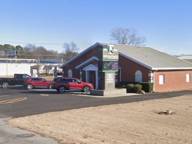 Brick church with a gabled roof and a sign for Refuge Church; cars parked in the front lot on a clear day.