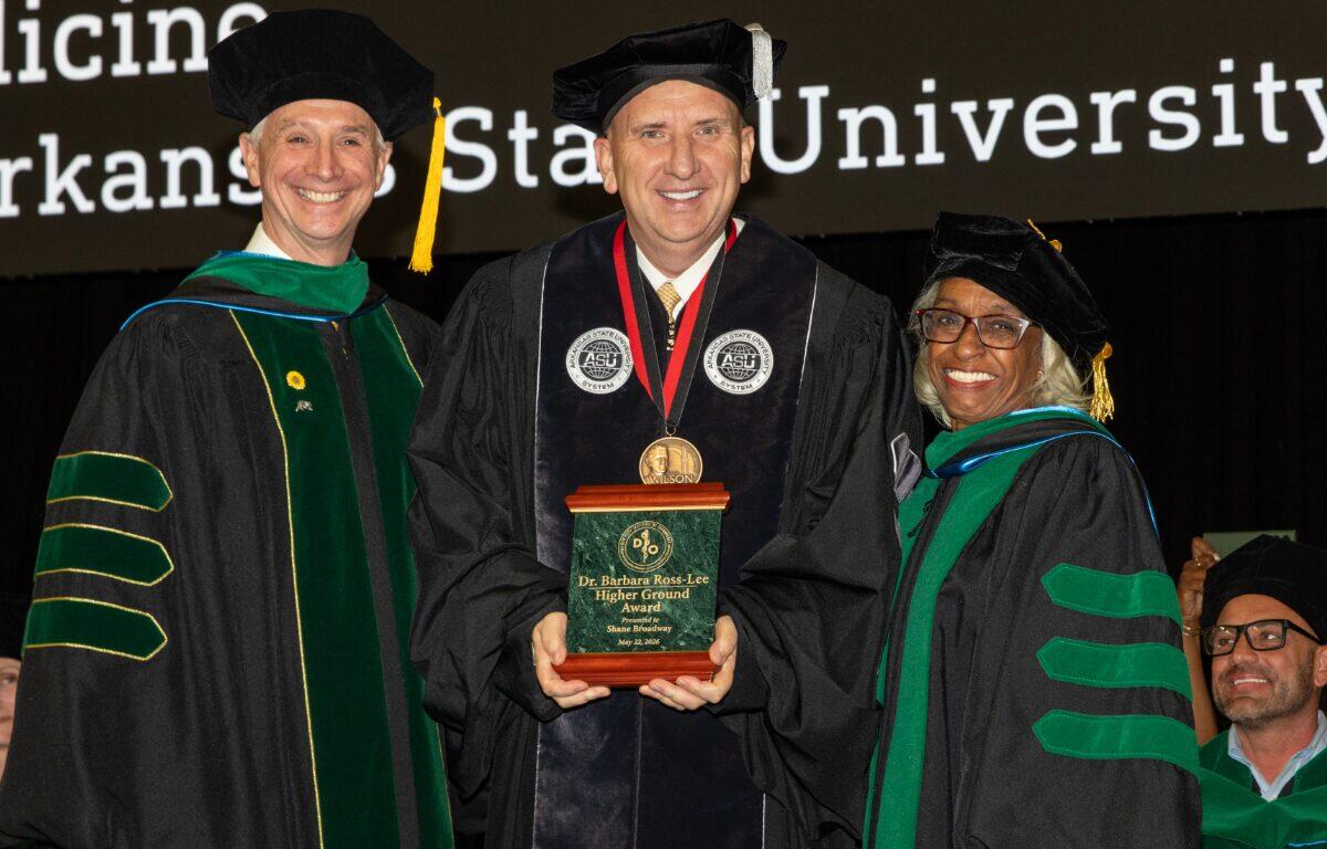 Three graduates in caps and gowns on stage; center recipient holds a plaque reading 'Higher Ground Award'.