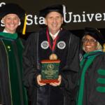 Three graduates in caps and gowns on stage; center recipient holds a plaque reading 'Higher Ground Award'.