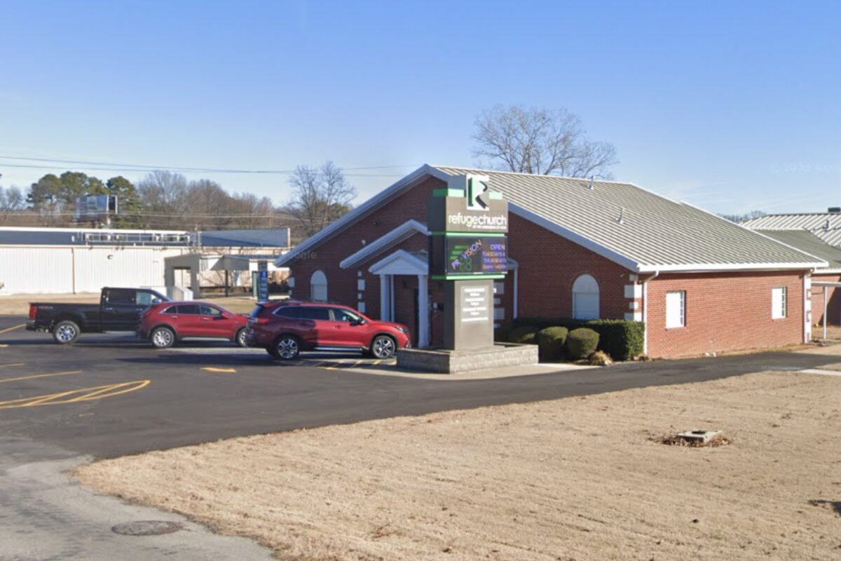 Brick church with a gabled roof and a sign for Refuge Church; cars parked in the front lot on a clear day.