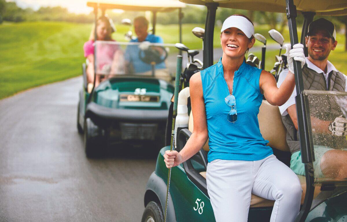 Smiling woman in a blue sleeveless golf shirt sits in a golf cart on a sunny course, with teammates in a second cart behind her.