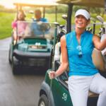 Smiling woman in a blue sleeveless golf shirt sits in a golf cart on a sunny course, with teammates in a second cart behind her.