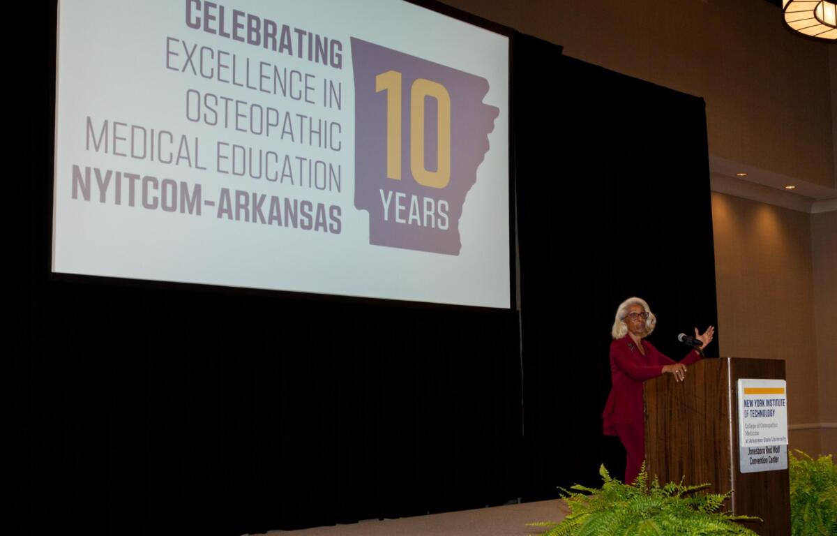 Speaker at a podium on a stage with a large screen showing a 10-year anniversary slide for NYITCOM-Arkansas Osteopathic Medical Education (celebrating 10 years) with a purple state silhouette in the graphic.