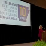 Speaker at a podium on a stage with a large screen showing a 10-year anniversary slide for NYITCOM-Arkansas Osteopathic Medical Education (celebrating 10 years) with a purple state silhouette in the graphic.