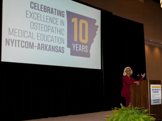 Speaker at a podium on a stage with a large screen showing a 10-year anniversary slide for NYITCOM-Arkansas Osteopathic Medical Education (celebrating 10 years) with a purple state silhouette in the graphic.
