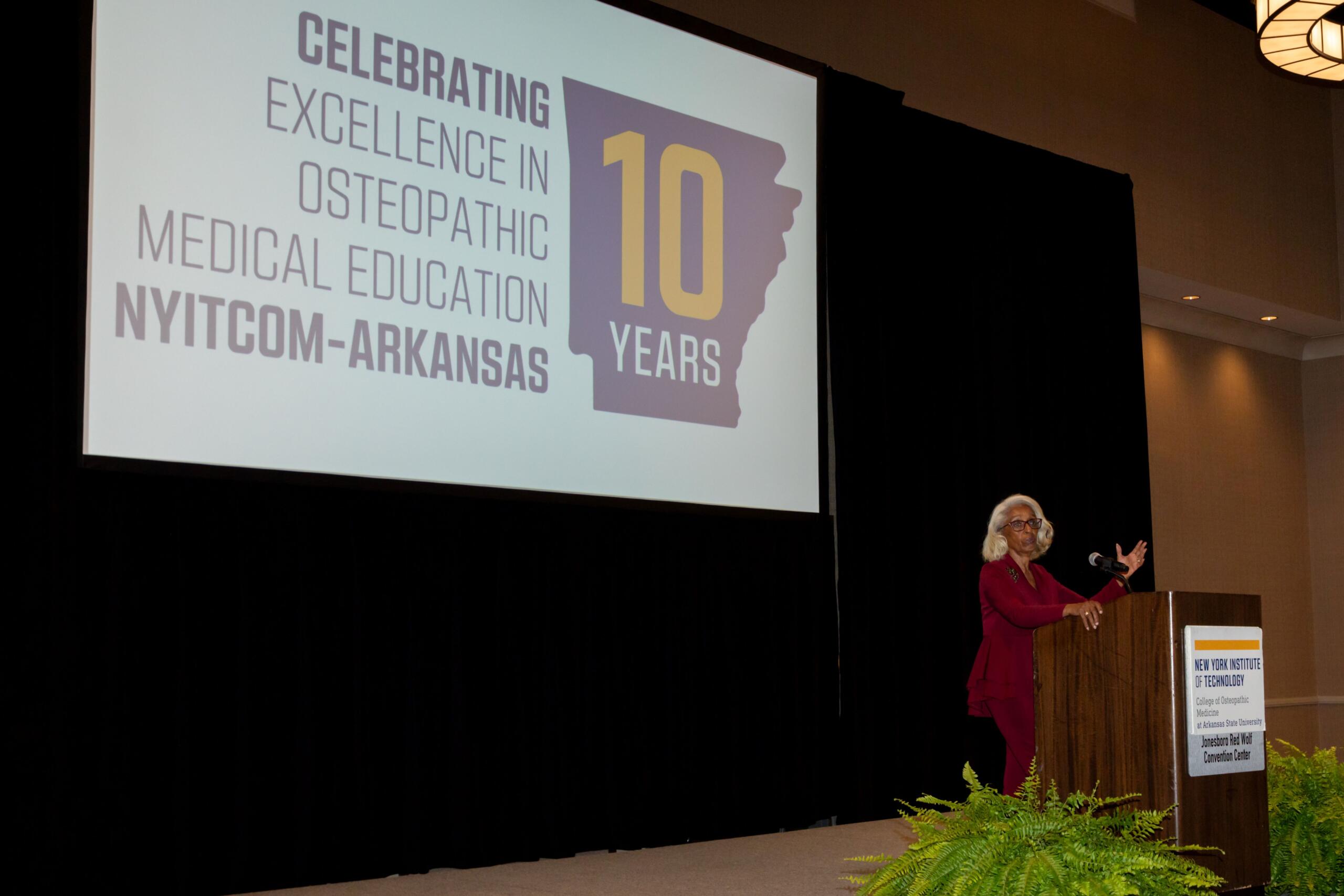 Speaker at a podium on a stage with a large screen showing a 10-year anniversary slide for NYITCOM-Arkansas Osteopathic Medical Education (celebrating 10 years) with a purple state silhouette in the graphic.