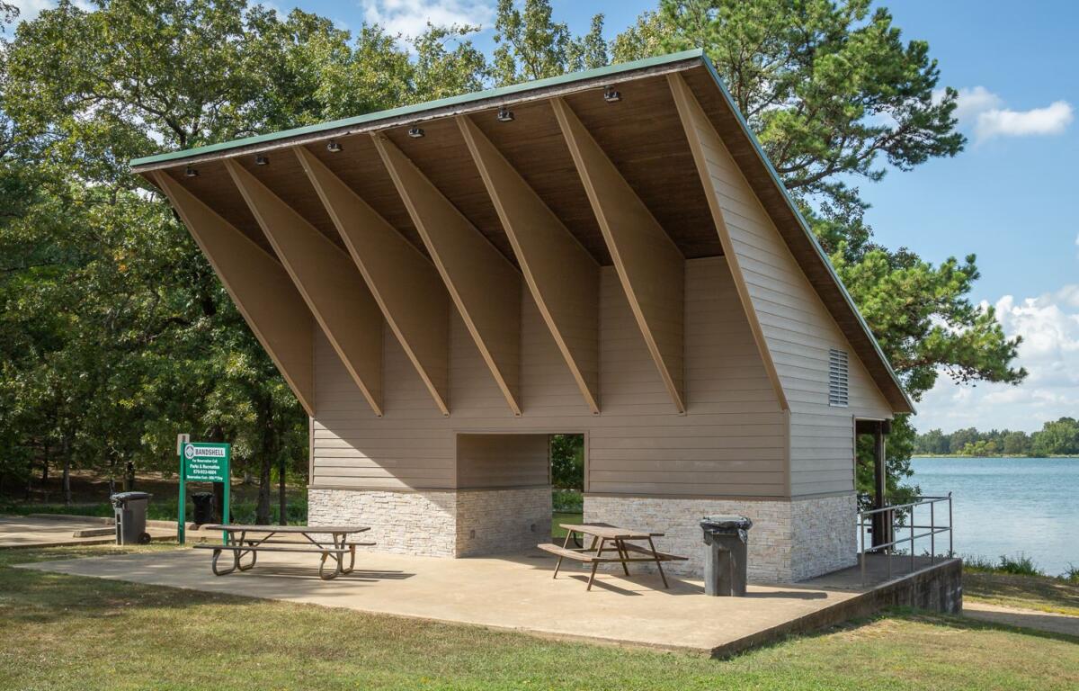 Park pavilion by a lake with a steeply pitched roof, beige siding, and a stone base; two picnic tables on the concrete pad.
