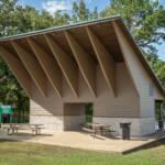 Park pavilion by a lake with a steeply pitched roof, beige siding, and a stone base; two picnic tables on the concrete pad.