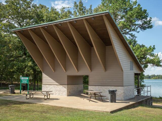 Park pavilion by a lake with a steeply pitched roof, beige siding, and a stone base; two picnic tables on the concrete pad.