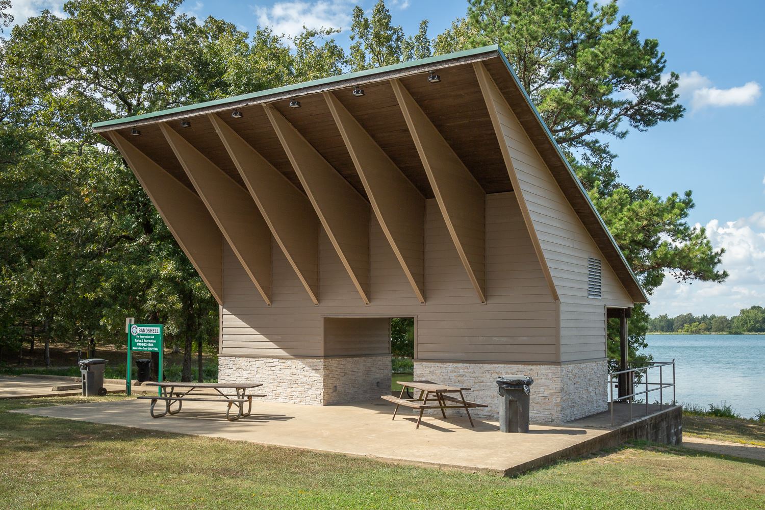 Park pavilion by a lake with a steeply pitched roof, beige siding, and a stone base; two picnic tables on the concrete pad.