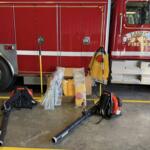 Red Lake City fire truck side with equipment on the floor in front, hoses, and tool bags scattered nearby inside a fire station bay.