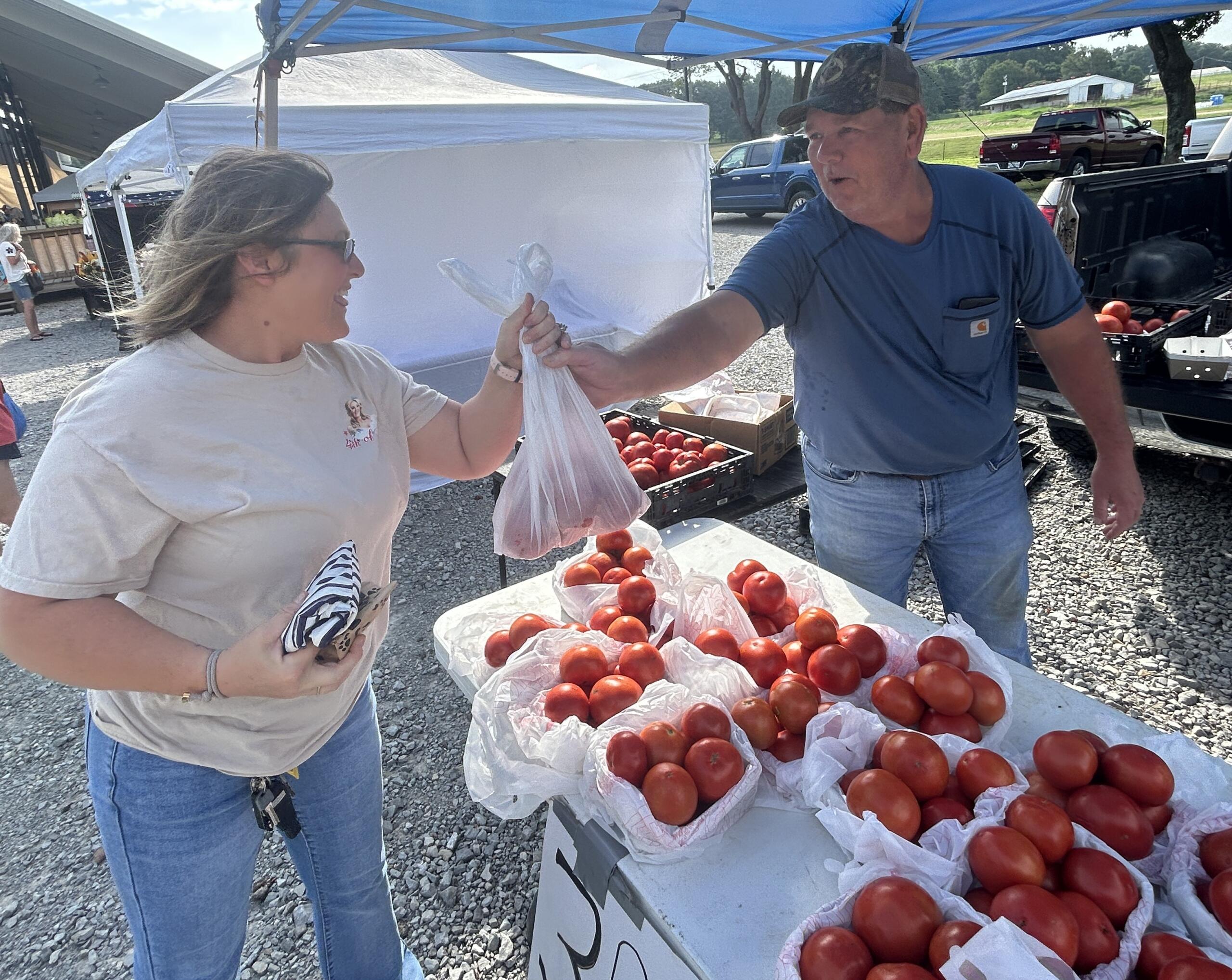 Two people at an outdoor market exchanging a plastic bag of tomatoes, with crates of fresh tomatoes on tables nearby.