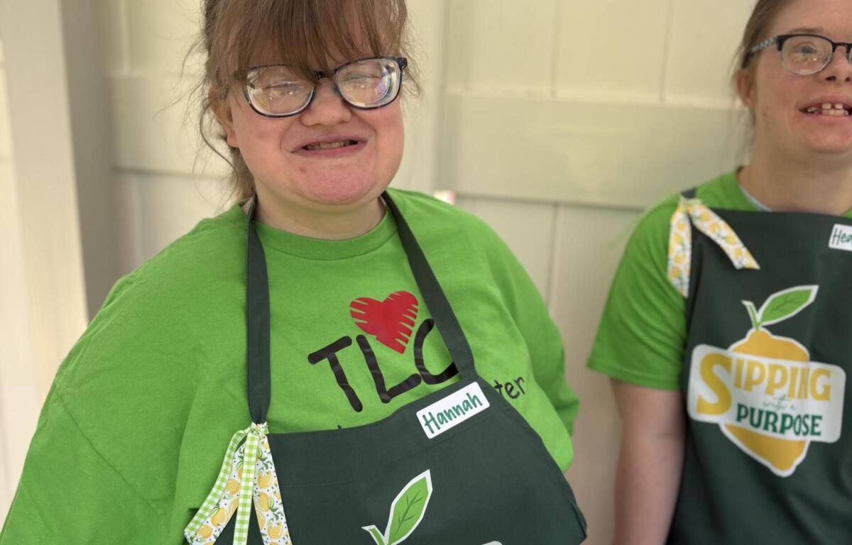 Two smiling women wearing green shirts and dark aprons at a booth, name tags read Hannah and another person, logo 'Sipping with a Purpose' on apron.