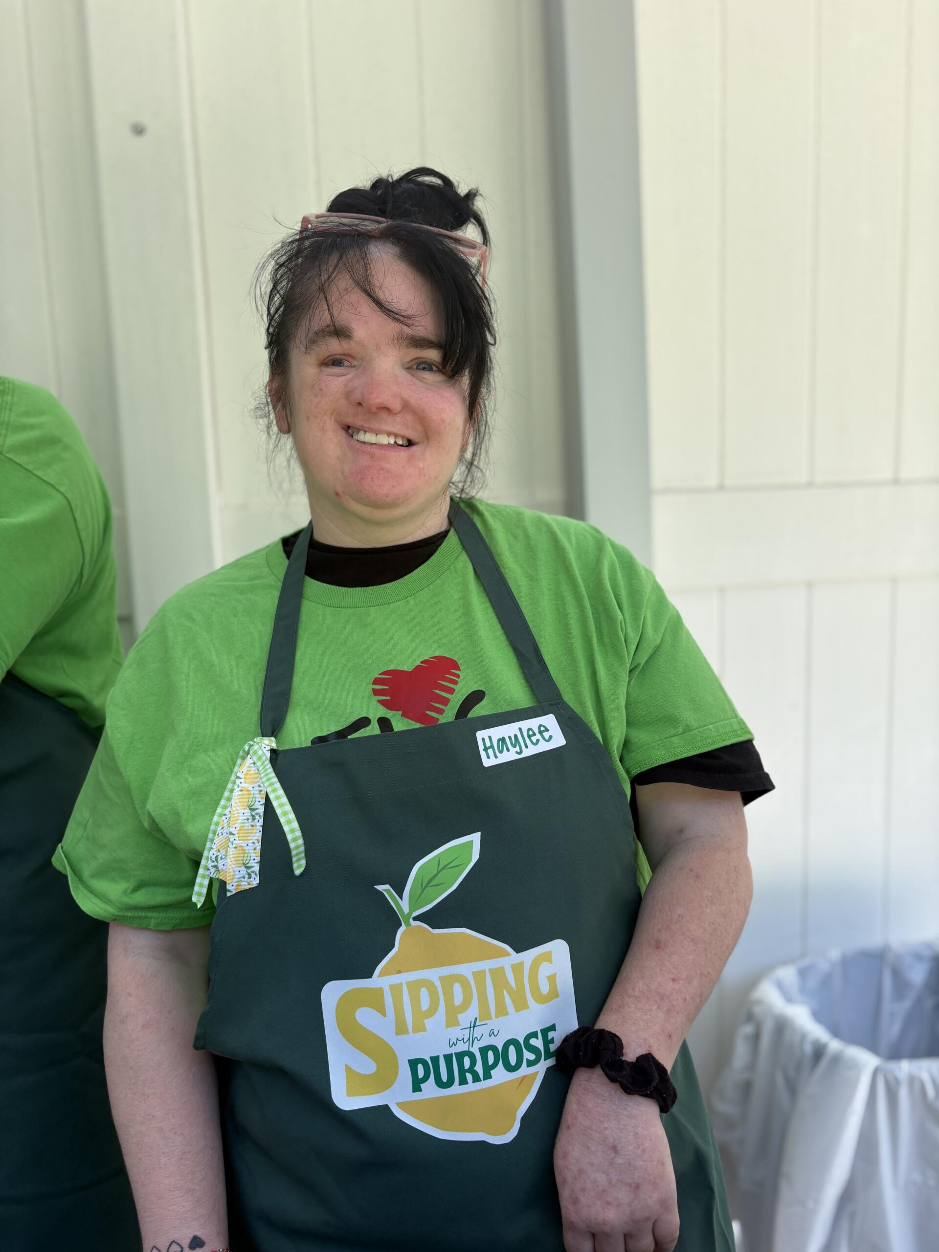 Smiling woman in a green shirt and dark apron with a name tag reading 'Haylee', sunglasses on her head, outdoors near a light wall.