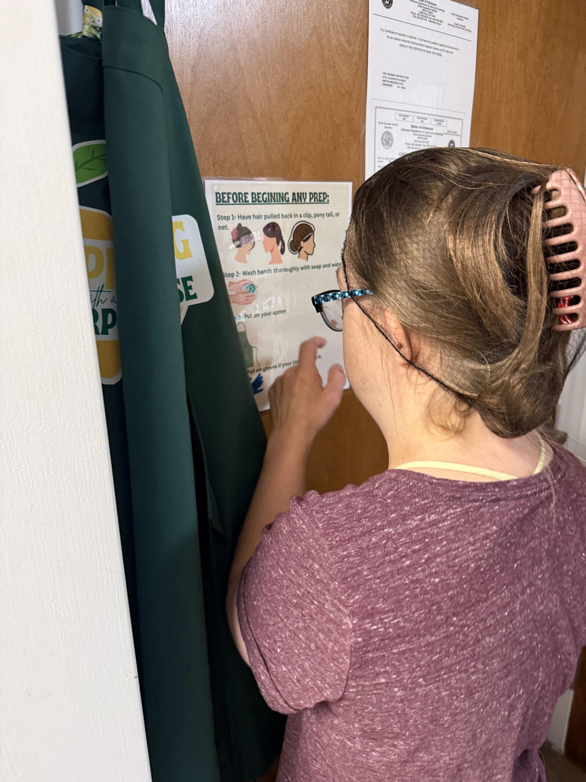 Back view of a person wearing a purple shirt and blue glasses, brushing or adjusting hair near a door with prep instruction posters.