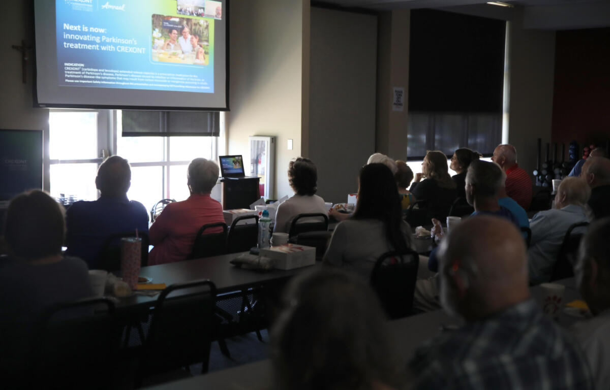 Audience seated in a conference room watching a projector screen displaying a presentation at the front of the room.