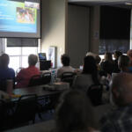 Audience seated in a conference room watching a projector screen displaying a presentation at the front of the room.