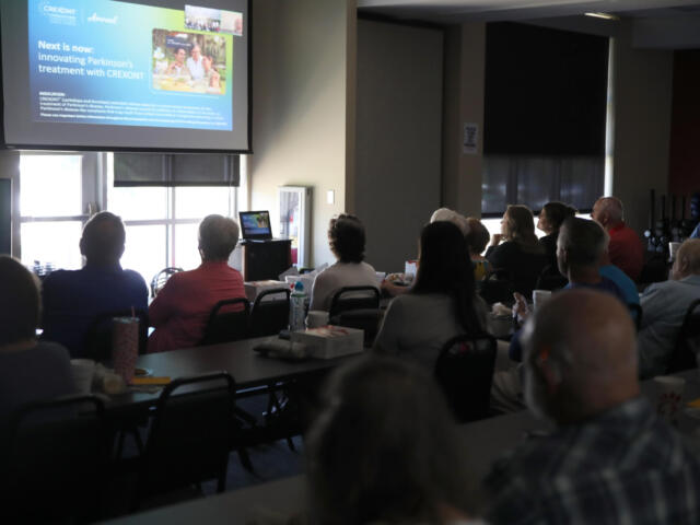 Audience seated in a conference room watching a projector screen displaying a presentation at the front of the room.