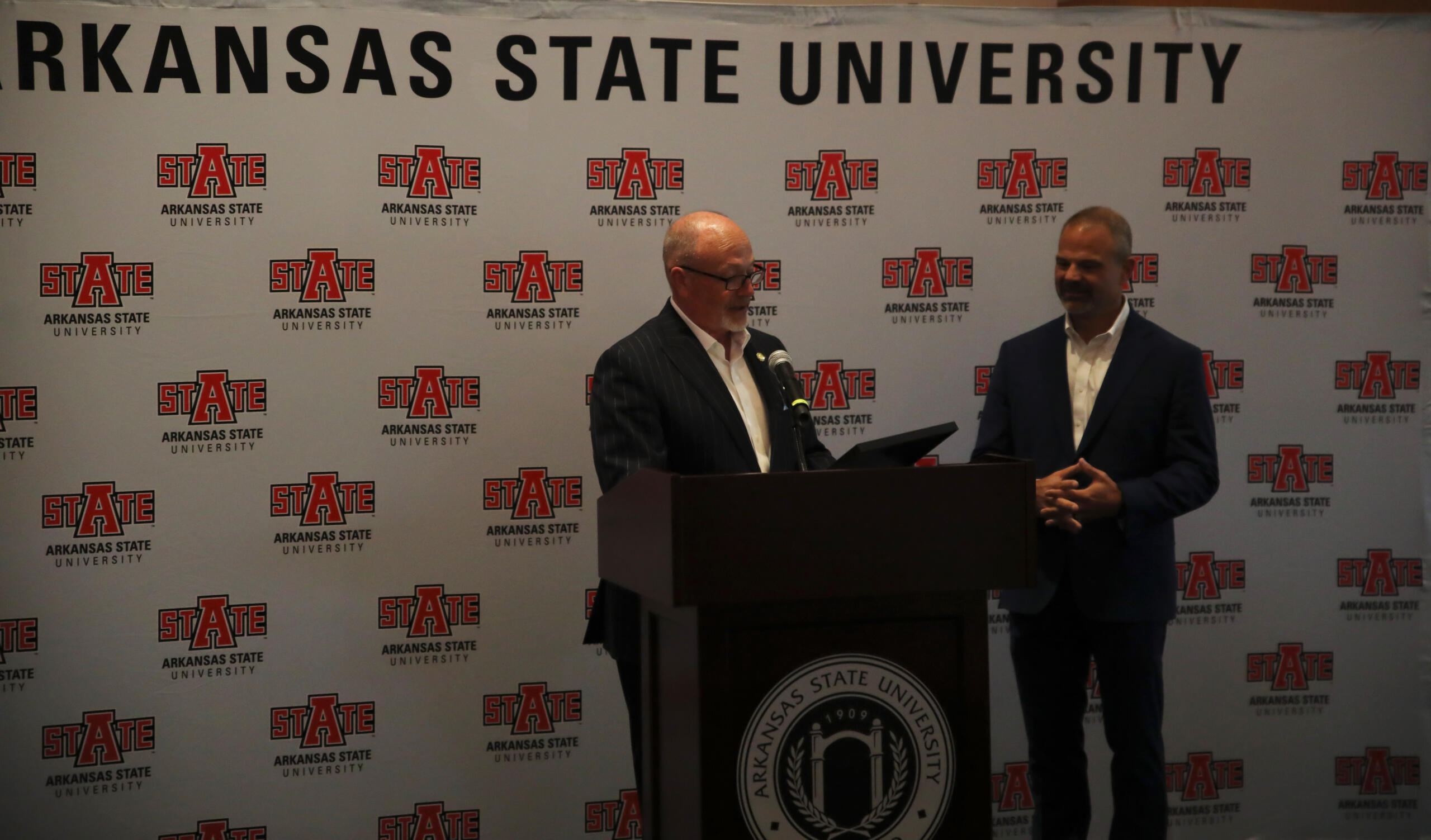 Two men in suits at a podium during a press event against an Arkansas State University backdrop.