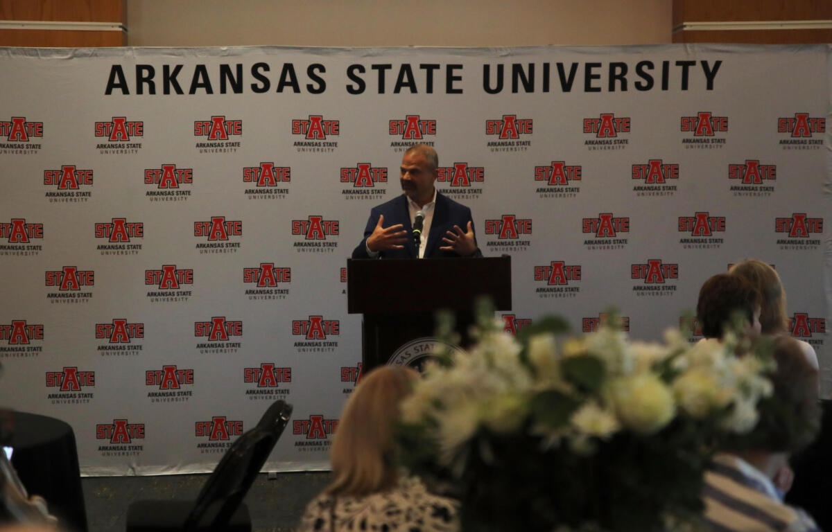 Speaker at a podium giving a talk in front of a backdrop covered with Arkansas State University logos and banners.