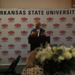 Speaker at a podium giving a talk in front of a backdrop covered with Arkansas State University logos and banners.