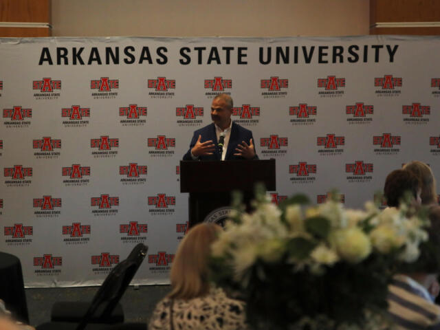 Speaker at a podium giving a talk in front of a backdrop covered with Arkansas State University logos and banners.