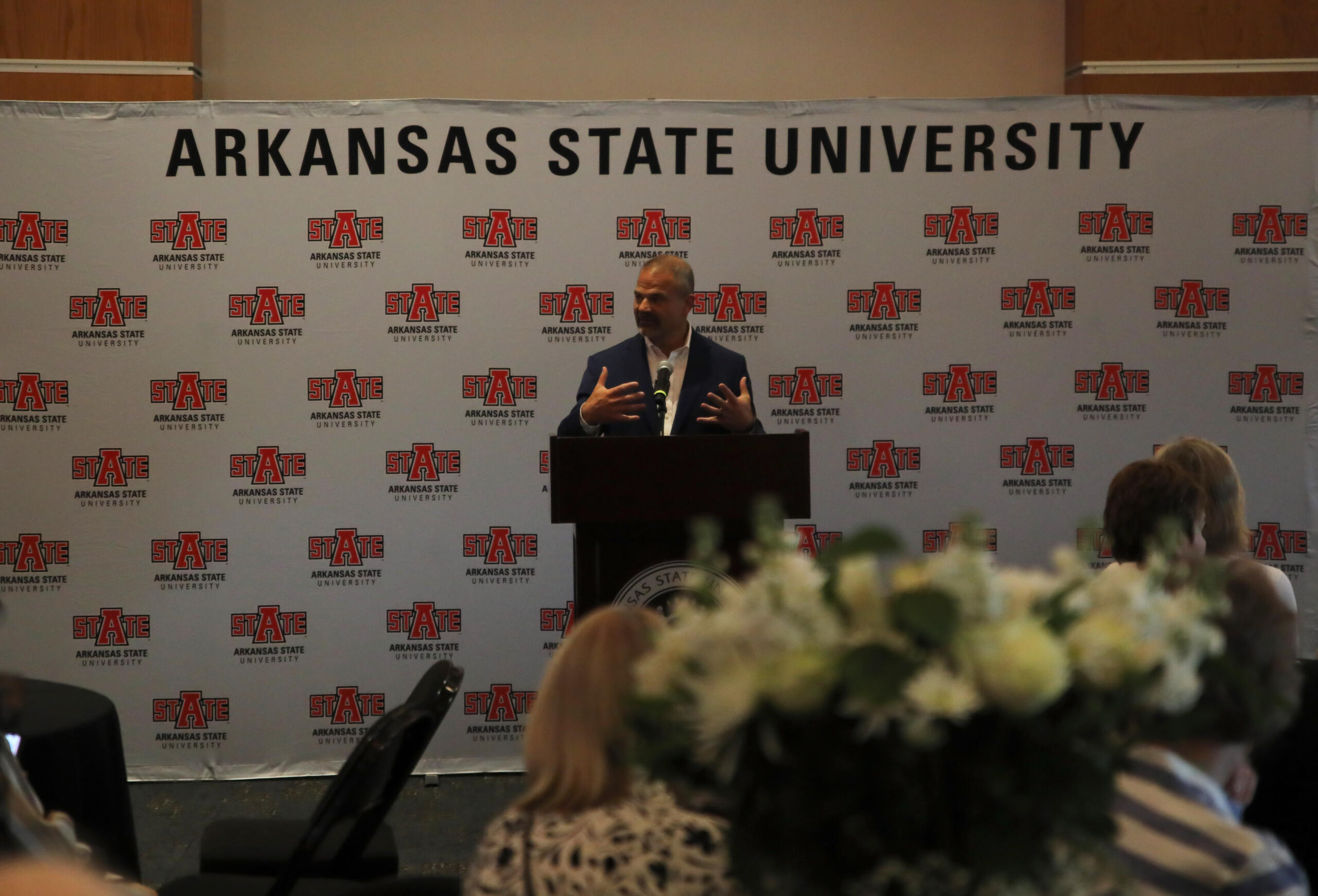 Speaker at a podium giving a talk in front of a backdrop covered with Arkansas State University logos and banners.