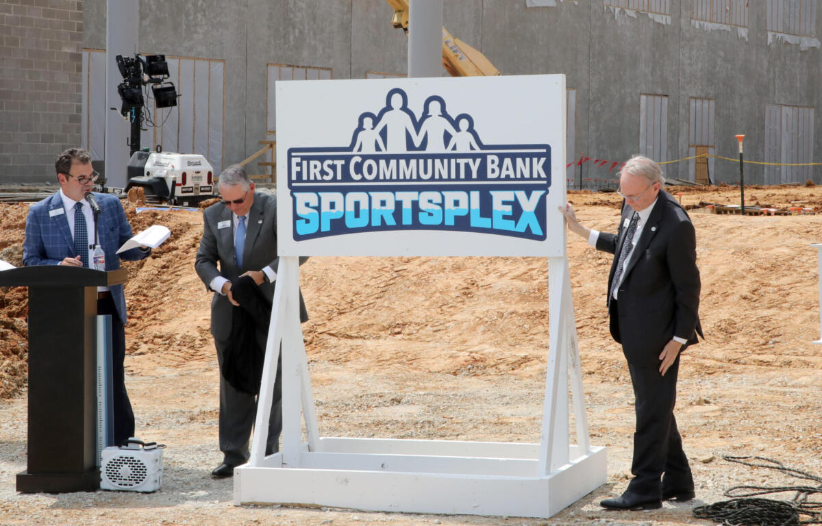 Three men in suits at a groundbreaking ceremony beside a 'First Community Bank Sportsplex' sign on a construction site.