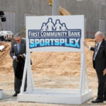 Three men in suits at a groundbreaking ceremony beside a 'First Community Bank Sportsplex' sign on a construction site.