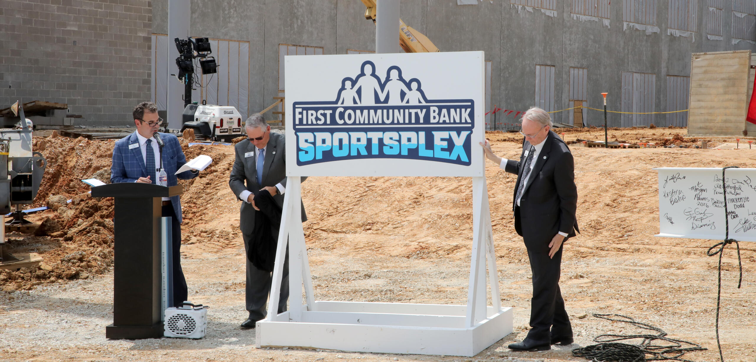 Three men in suits at a groundbreaking ceremony beside a 'First Community Bank Sportsplex' sign on a construction site.