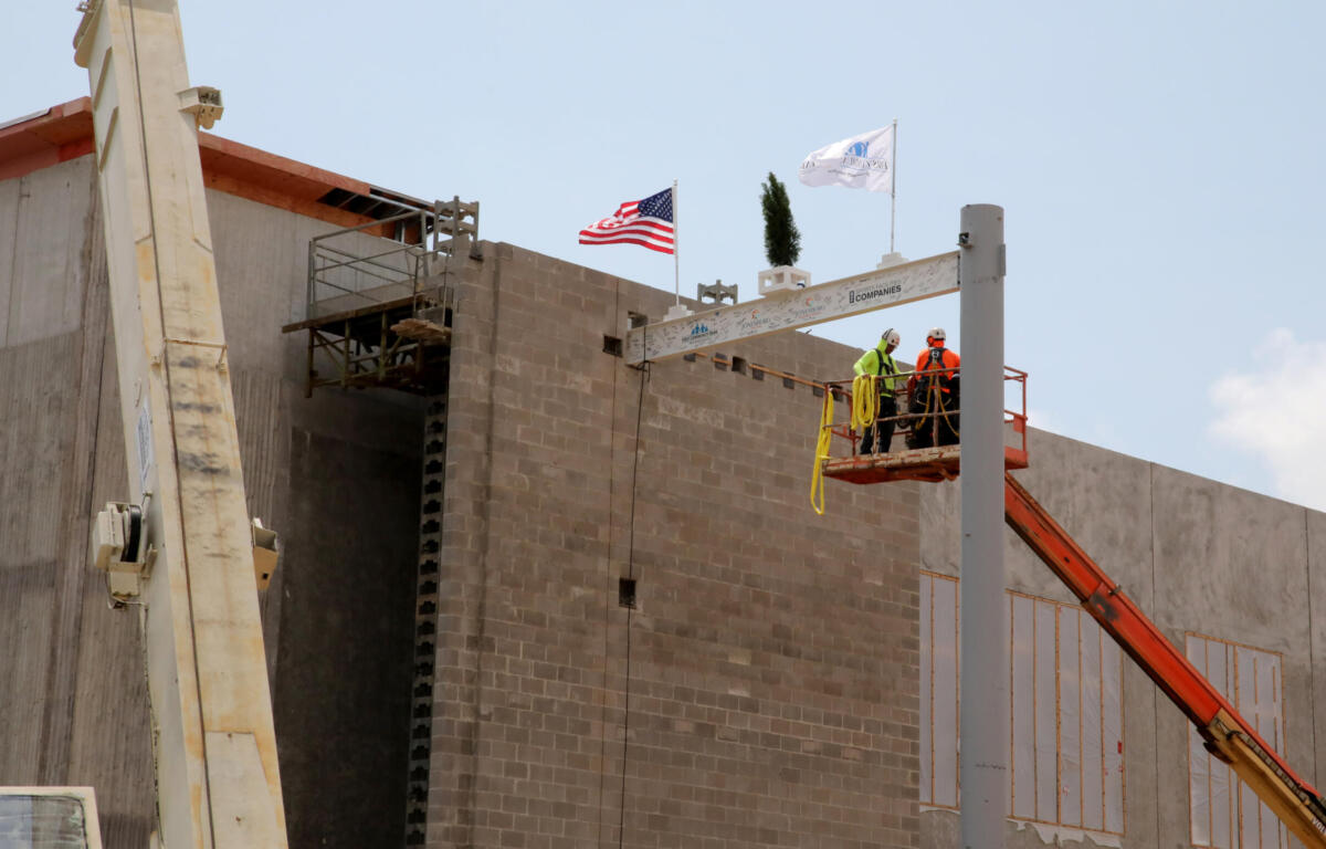 Two construction workers on a raised platform at a building site near an American flag under a blue sky.