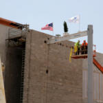 Two construction workers on a raised platform at a building site near an American flag under a blue sky.