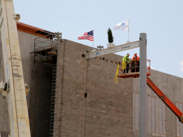 Two construction workers on a raised platform at a building site near an American flag under a blue sky.