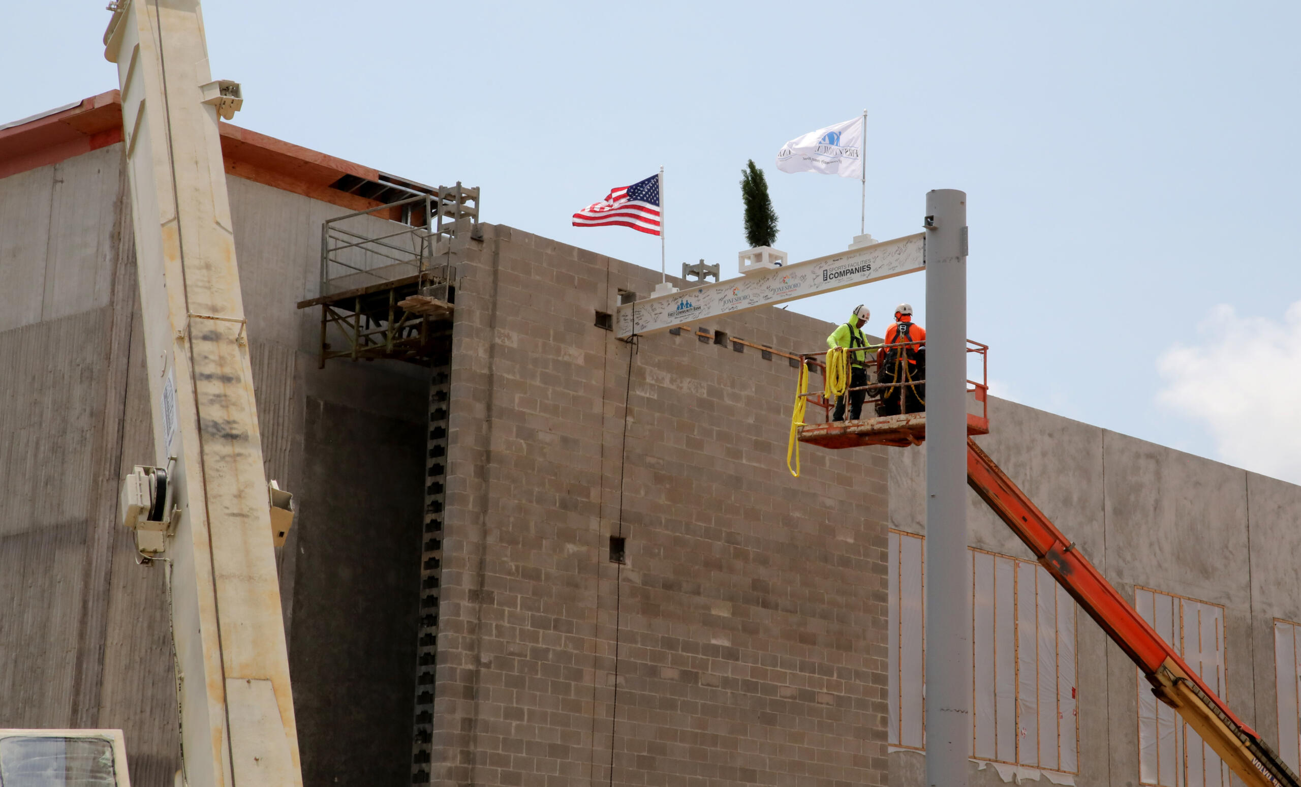 Two construction workers on a raised platform at a building site near an American flag under a blue sky.