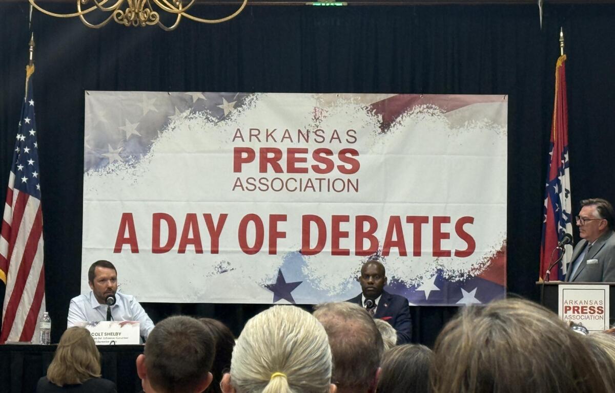 Panel of speakers on a stage at an Arkansas Press Association event, with flags on either side and a banner reading 'A DAY OF DEBATES' in the background.