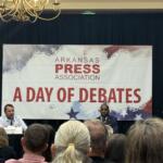 Panel of speakers on a stage at an Arkansas Press Association event, with flags on either side and a banner reading 'A DAY OF DEBATES' in the background.