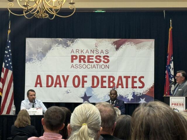 Panel of speakers on a stage at an Arkansas Press Association event, with flags on either side and a banner reading 'A DAY OF DEBATES' in the background.