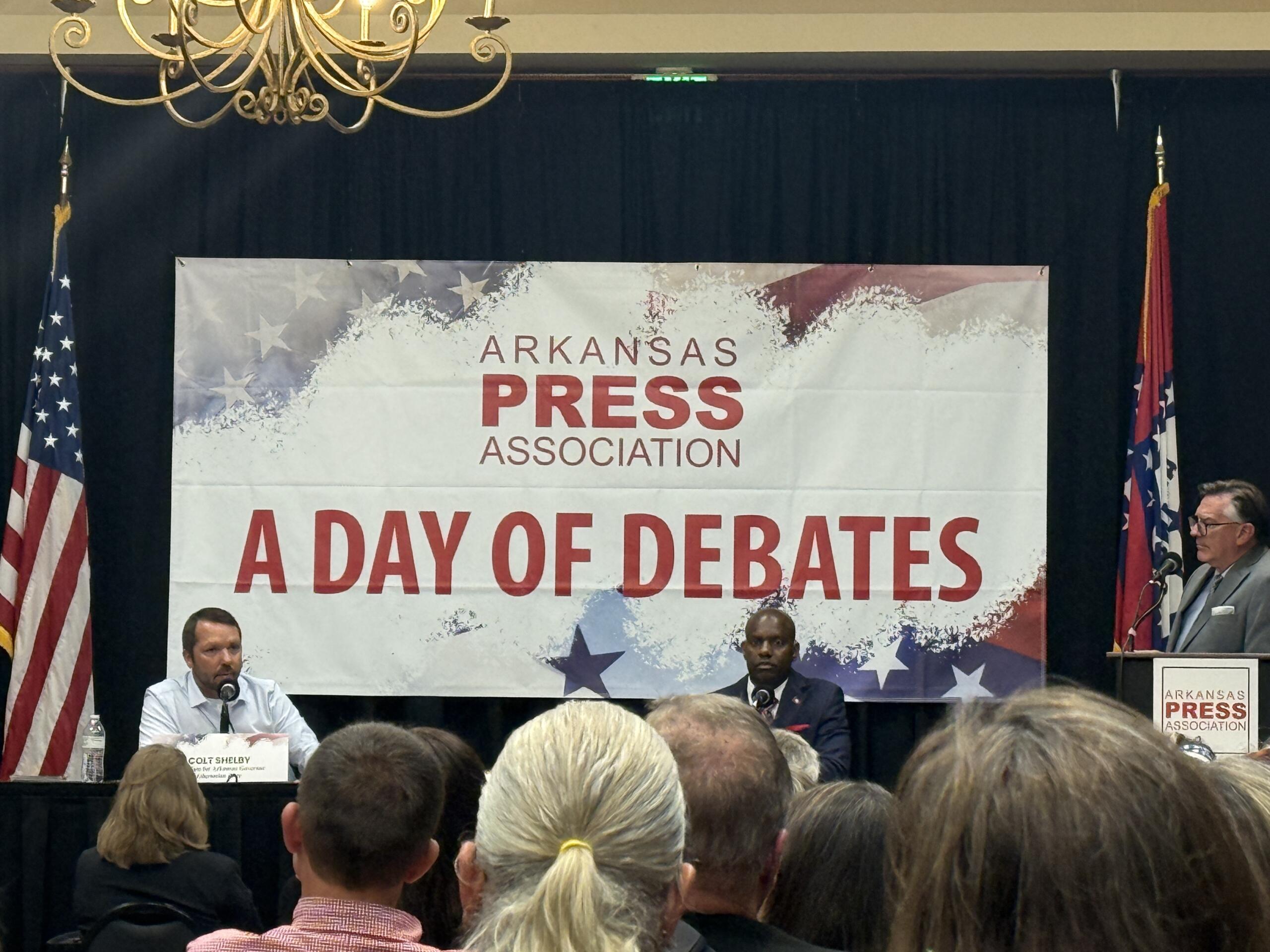 Panel of speakers on a stage at an Arkansas Press Association event, with flags on either side and a banner reading 'A DAY OF DEBATES' in the background.