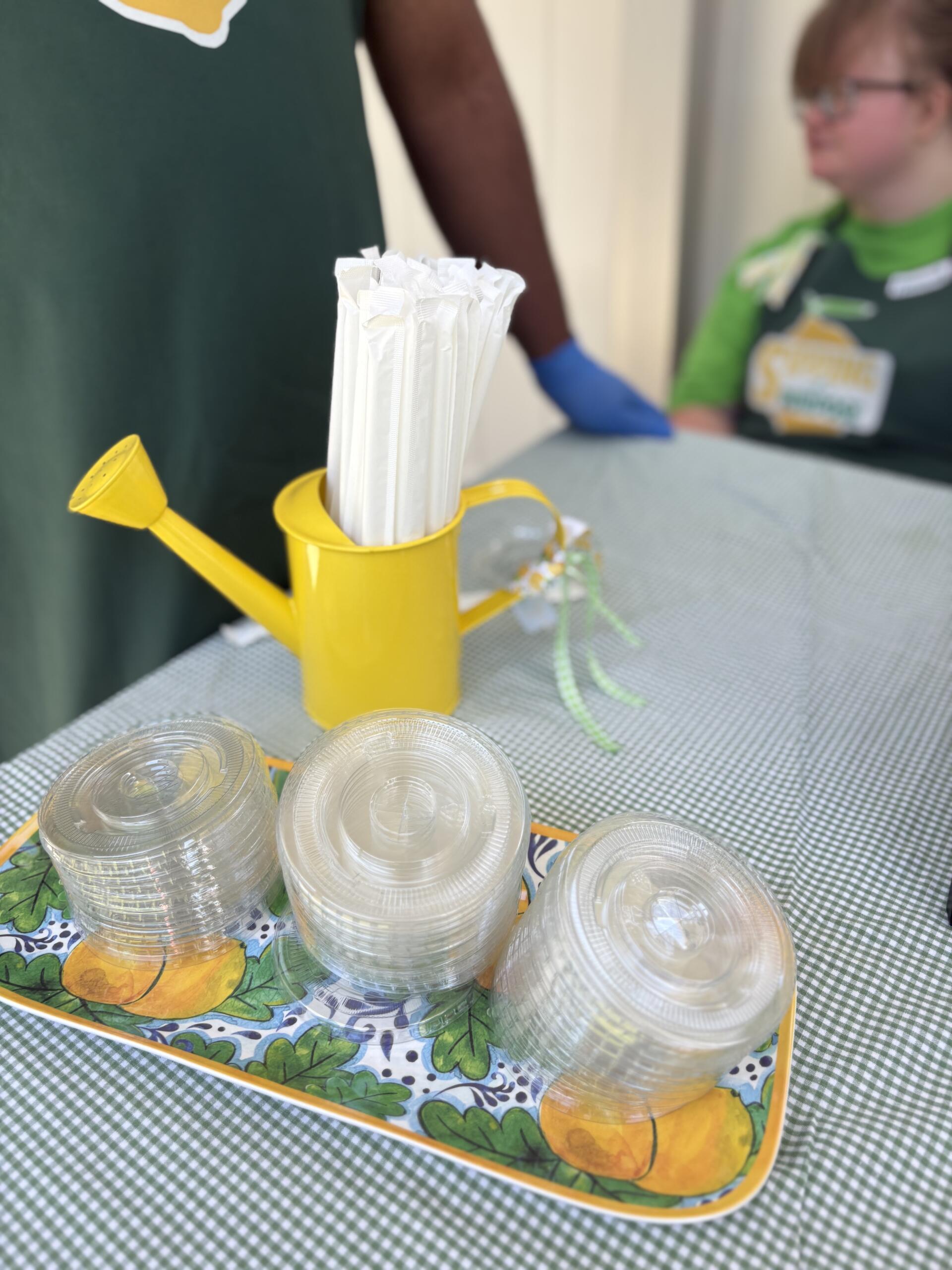Yellow watering can holding white folded napkins in a lemon-patterned tray, on a green-and-white checkered tablecloth, with clear plastic cups nearby and blurred people in the background.