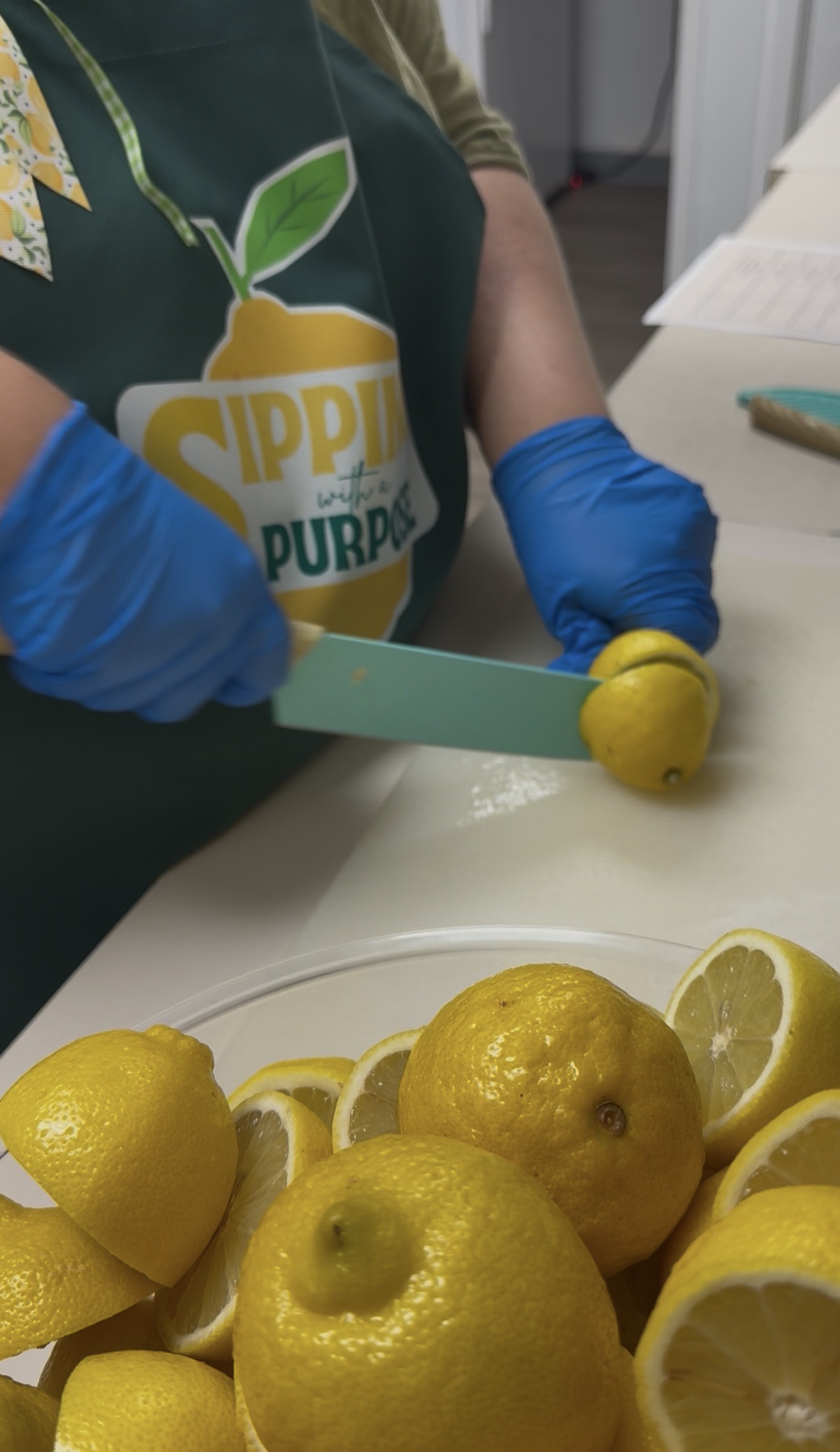 Person wearing blue gloves and a dark green apron slices a lemon with a knife at a kitchen prep station.