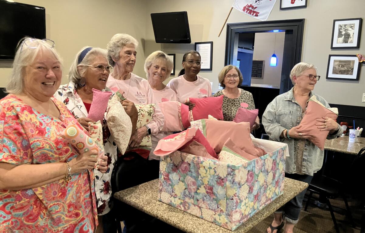 Smiling group of women holding pink gift bags at a party, standing around a counter with colorful gift wrap in a cozy room.