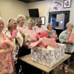 Smiling group of women holding pink gift bags at a party, standing around a counter with colorful gift wrap in a cozy room.