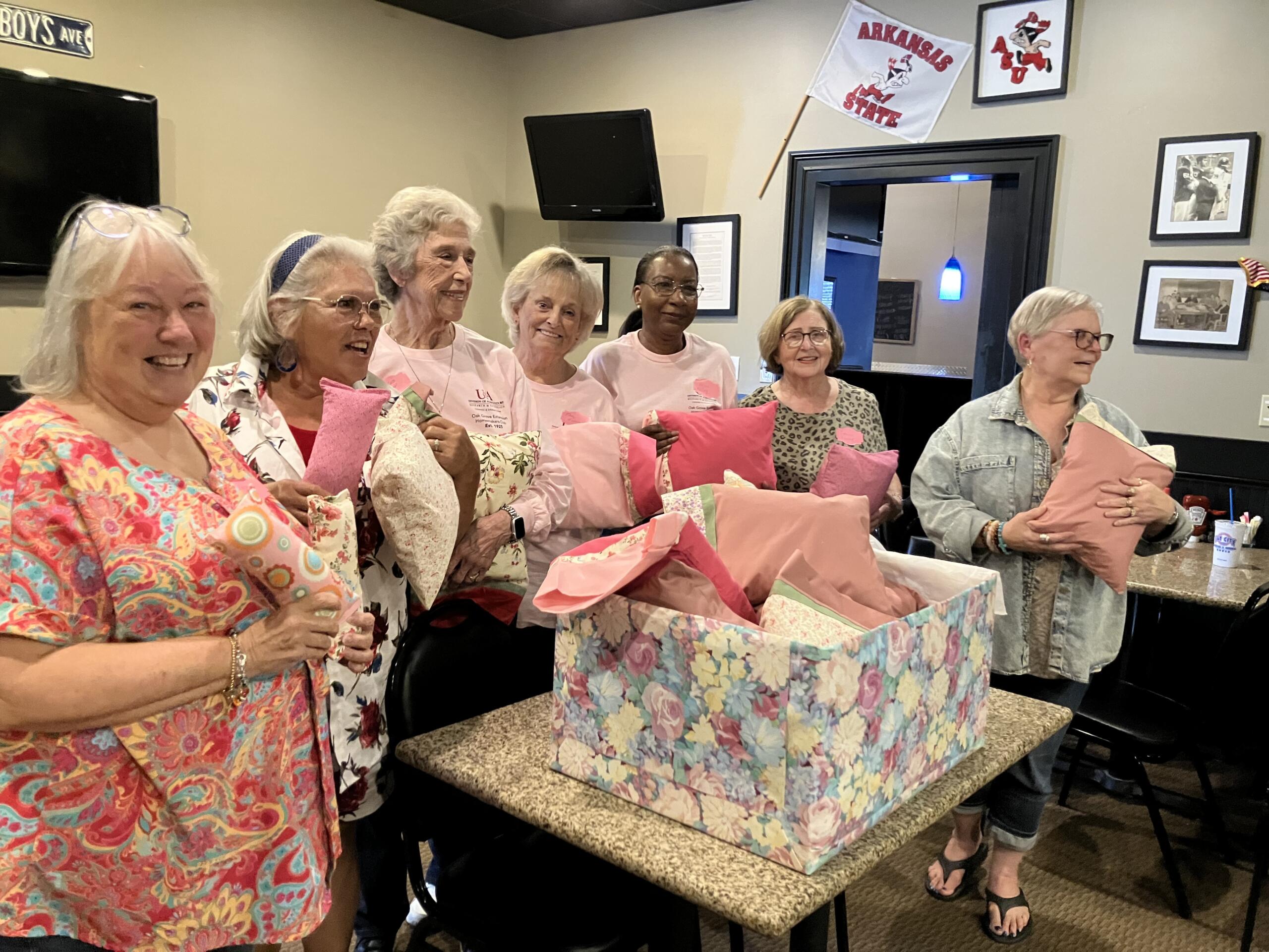 Smiling group of women holding pink gift bags at a party, standing around a counter with colorful gift wrap in a cozy room.
