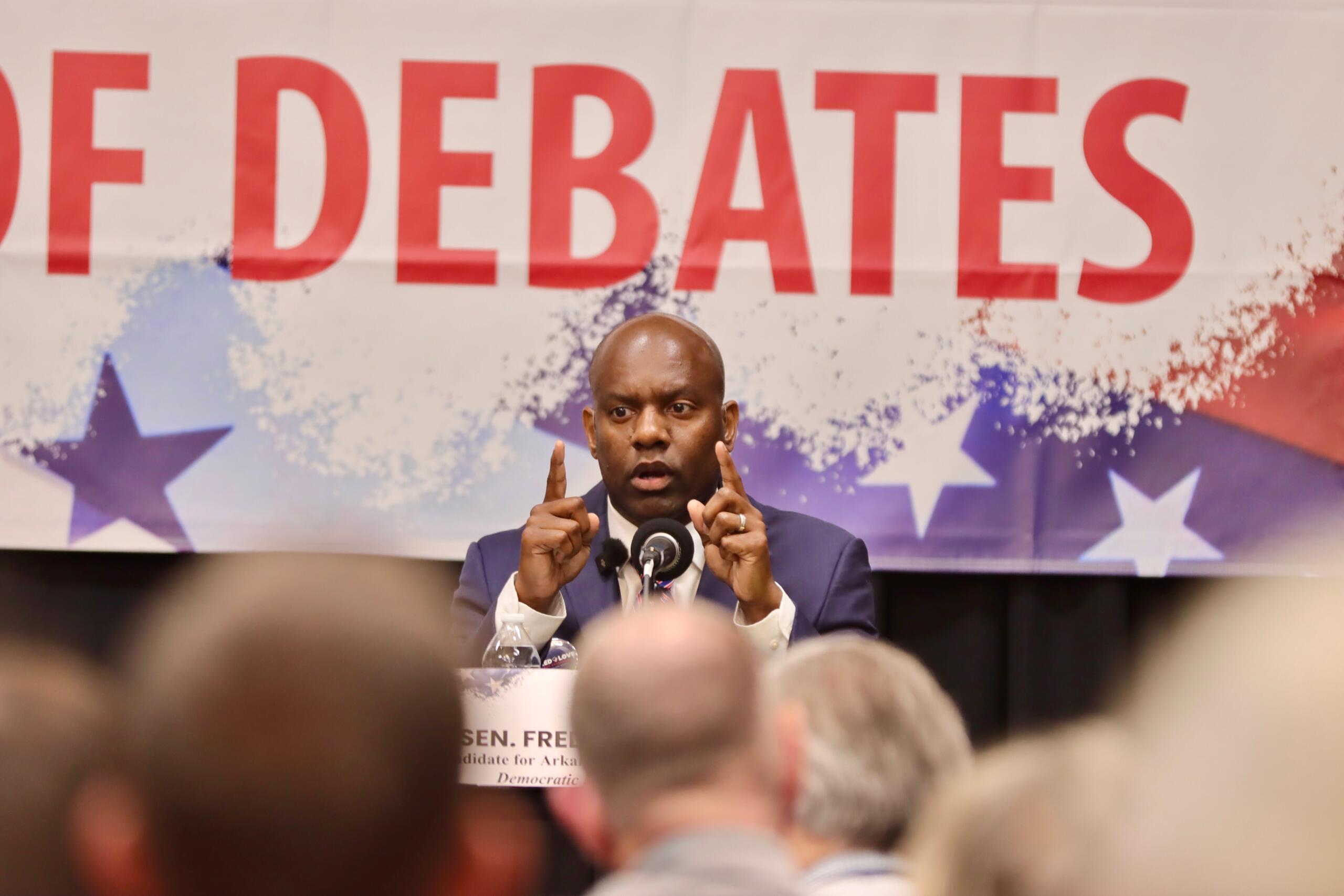 Male speaker at a political debate podium gesturing with both hands, with a large 'DEBATES' banner in the background