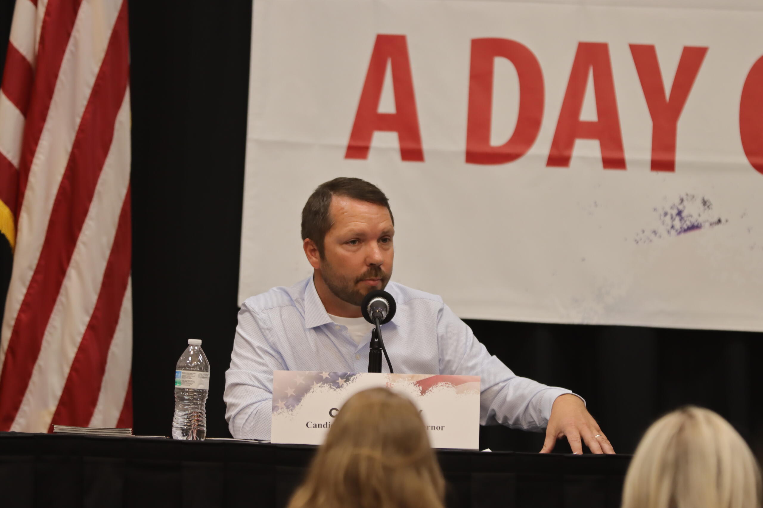 Man in a light blue shirt speaks into a microphone at a panel on a stage, with an American flag on the left and a large banner behind him.