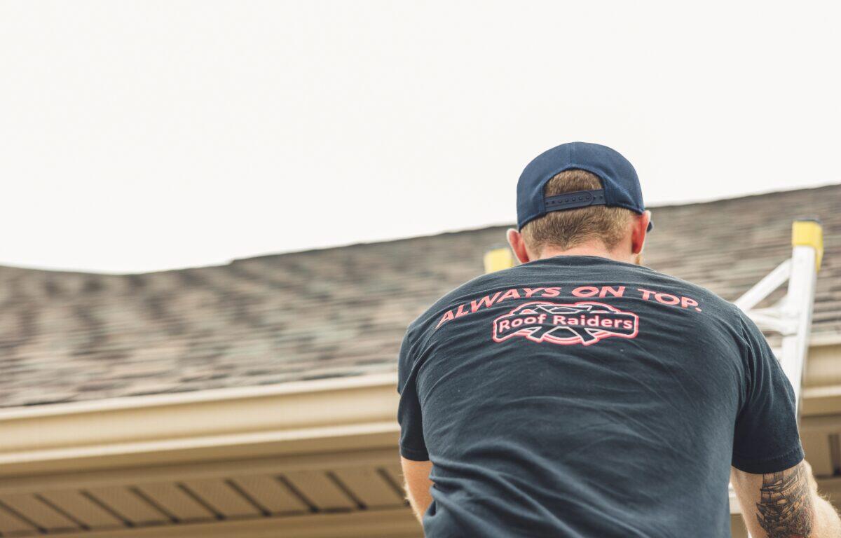 Man on a ladder at a roofline, seen from behind, wearing a dark T-shirt that reads 'Always on Top' with a Roof Raiders logo.